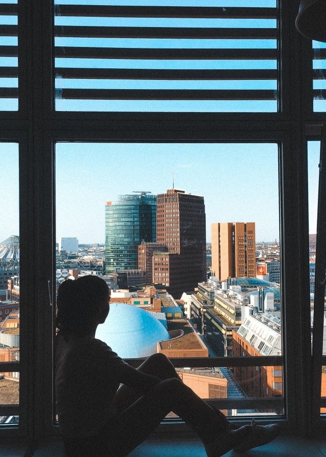 A girl is sitting inside of her window and looking out at the city.