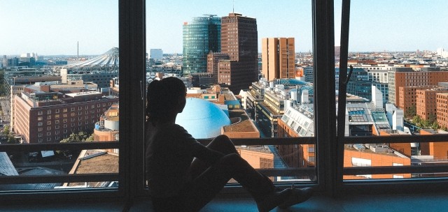 A girl is sitting inside of her window and looking out at the city.